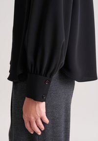 Blusa nera con maniche lunghe e a sbuffo, dotata di polsini rotondi con due bottoni marroni, abbinata a un tessuto grigio testurizzato sottostante.
