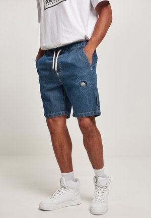 Short en jean avec une ceinture élastique, un cordon de serrage, des poches latérales et un logo patch sur la cuisse gauche. Porté avec un t-shirt blanc et des baskets.