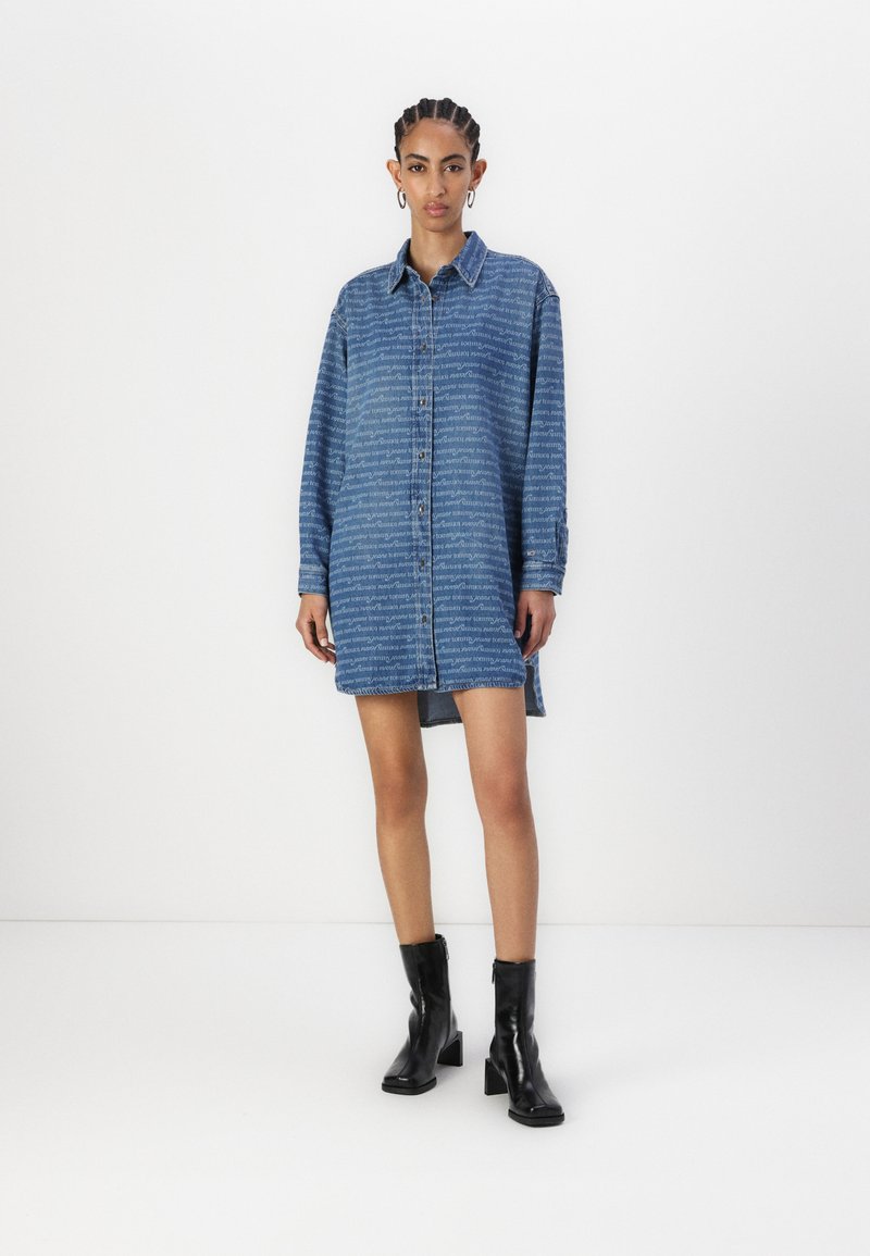 Blaues Denim-Hemdkleid mit Knopfleiste, langen Ärmeln und seitlichen Schlitzen. Verfügt über ein wiederholtes Textmuster und wird mit schwarzen Knöchels Stiefeln kombiniert.