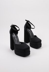 Tacones de plataforma negros con puntera cuadrada, tacón de bloque grueso y correa en el tobillo. Fabricados con un tejido suave y un acabado mate.