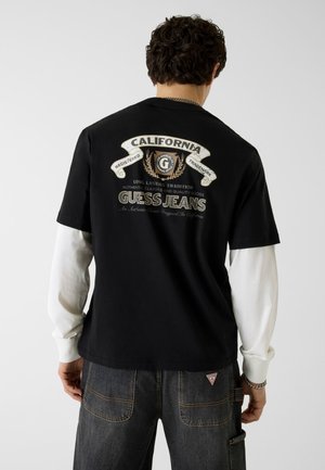 Mann von hinten, trägt ein schwarzes Guess Jeans T-Shirt über weißen langen Ärmeln und dunkle Jeans, Logo und Aufschrift „California“ sind auf dem Rücken des Shirts sichtbar.