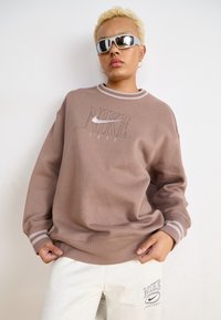 Felpa Nike marrone con logo e anno "1972", caratterizzata da polsini a coste e scollo rotondo, abbinata a pantaloni della tuta chiari.