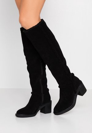 Femme portant des bottes en suède noir montant jusqu'aux genoux avec des talons carrés et des détails de couture en zigzag, debout devant un fond clair uni.