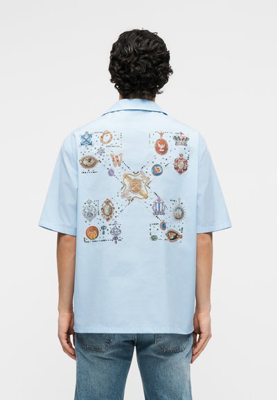 Άτομο που φοράει ανοιχτό μπλε κοντομάνικο πουκάμισο με πολύχρωμες, περίτεχνες τσέπες και διακοσμητικά που σχηματίζουν ένα σχήμα Χ στην πλάτη.