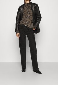 Kvinna klädd i en mörkblommig blus, svart läderjacka över axlarna, svarta rakbenade jeans och svarta klackboots.