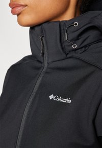 Czarna kurtka softshell z wysokim kołnierzem, zamkiem błyskawicznym, regulowanym kapturem oraz białym logo Columbia na lewej części klatki piersiowej.
