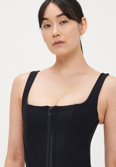 Černý overal na zip s hranatým výstřihem, širokými ramínky a přiléhavým střihem, s texturovanou tkaninou a nenápadným logem na zipu.