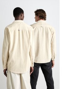 Dos personas están de espaldas, vistiendo camisas de corderoy de color beige claro con cuello y puños abotonados, mostrando una textura acanalada.