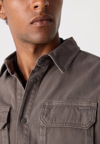 Camisa gris de botones con cuello, que cuenta con dos bolsillos en el pecho y un tejido texturizado con un sutil patrón a rayas.