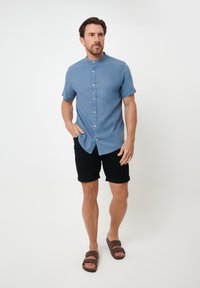 Lichtblauw kortärmige overhemd met kraag, door middel van knopen aan de voorkant, gecombineerd met opgerolde zwarte shorts en bruine sandalen. Neutrale achtergrond.
