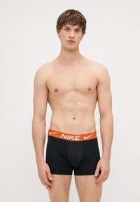 Μαύρα μποξέρ με πορτοκαλί λογότυπο Nike στην ελαστική μέση. Το ύφασμα φαίνεται λείο και ελαστικό, αγκαλιάζοντας στενά το σώμα.