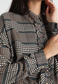 Camicia con bottoni dalla fantasia geometrica nei toni del marrone, crema e teal; presenta un colletto, maniche con risvolto e piccoli bottoni metallici.