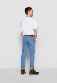 Világoskék farmernadrág, keskenyített fazonnal, hátsó zsebekkel és barna bőr folttal a derékrészen, fehér pólóval és barna csizmával kombinálva.