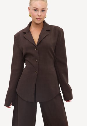 Mujer con cabello rubio, lleva pendientes dorados y un blazer marrón oscuro abotonado con pantalones anchos a juego sobre un fondo blanco liso.