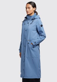 Abrigo impermeable azul claro con capucha, botones a presión en el frente, dos bolsillos laterales, puños ajustables y un parche negro con el logo en la manga.