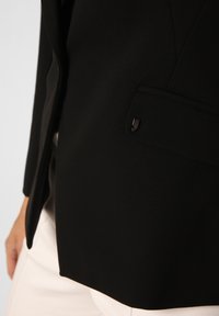 Schwarzer, taillierter Blazer mit glatter Textur, der eine Brusttasche und ein kleines Metallakzent am Kragen für Details aufweist.