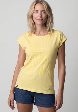 Blonde Frau mit welligem Haar trägt ein gelbes ärmelloses Shirt mit schwachem Text und dunkelblaue hochgekrempelte Jeansshorts, steht vor schlichtem grauem Hintergrund.