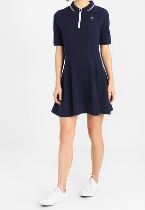 Femme portant une robe polo bleu marine à manches courtes avec des liserés blancs, associée à des baskets blanches, debout devant un fond blanc uni.