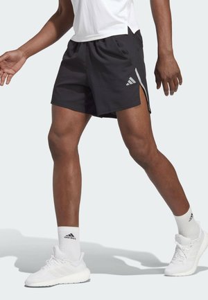 Osoba ubrana w czarne spodenki Adidas, białe skarpetki Adidas i białe buty sportowe Adidas, stojąca w pozie chodzącej na jednolitym tle.