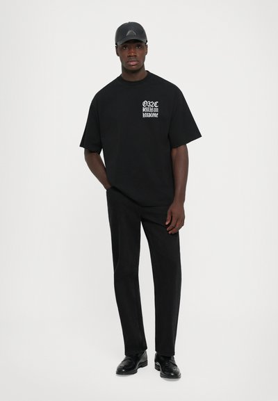 Jeune homme portant une casquette noire, un t-shirt noir ample avec du texte blanc, un pantalon noir et des chaussures noires, se tenant devant un fond blanc uni.