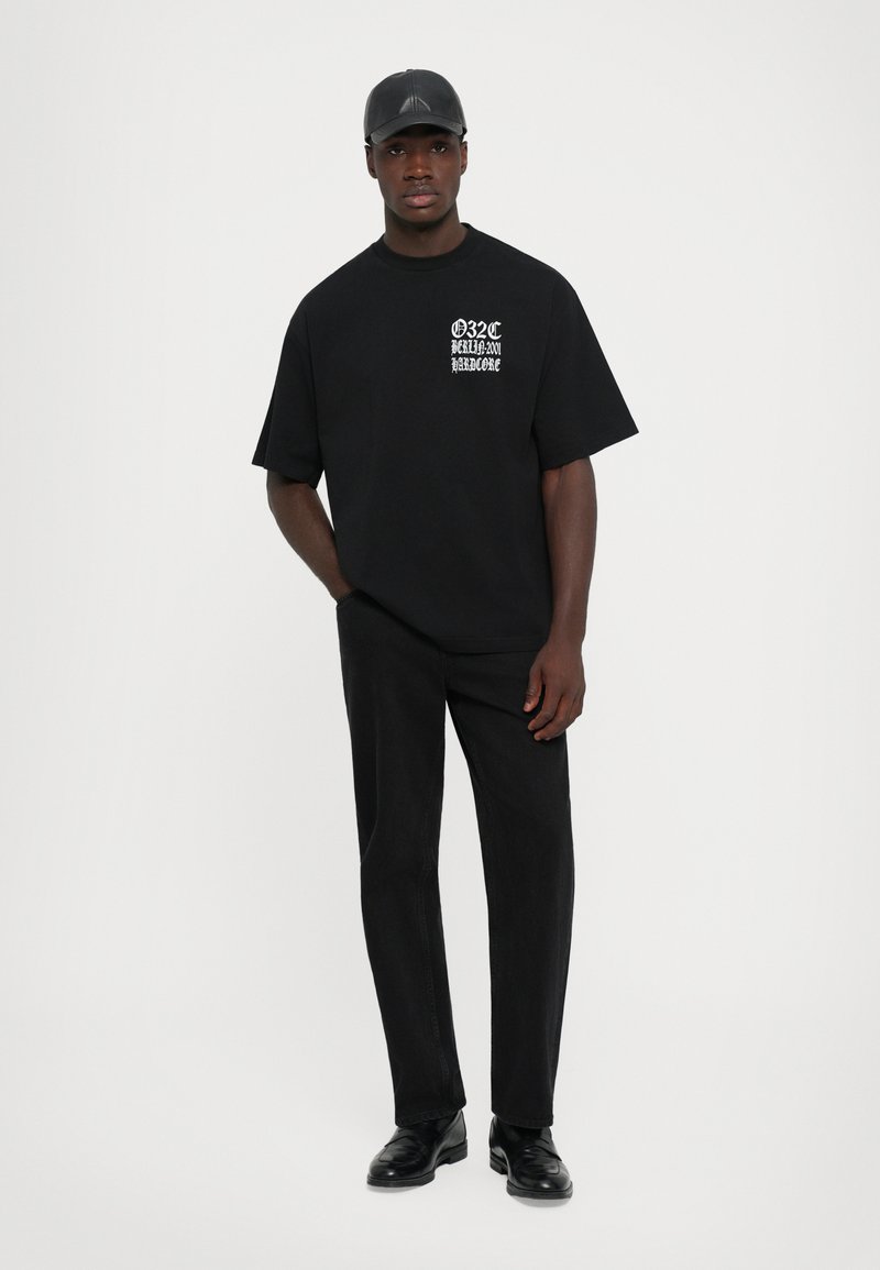 Young man wearing black cap, oversized black t-shirt with white text, black pants, and black shoes, standing against a plain white background.