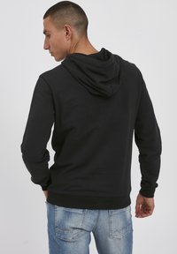 Svart hettegenser med en tettsittende design, teksturert stoff og justerbar hette. Sammen med lyseblå denimjeans. Utsikten er bakfra.