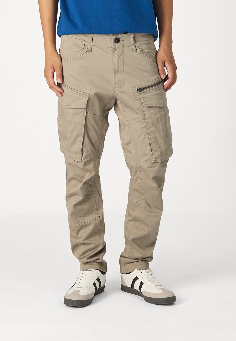 Pantalon cargo beige avec plusieurs poches, porté par une personne en chemise bleue et baskets blanches avec des bandes noires, debout sur fond blanc.