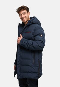 Donkerblauwe gewatteerde jas met capuchon, zijzakken, ritssluiting en rood logo op de borst. Gestructureerd, geïsoleerd materiaal met een losse pasvorm.