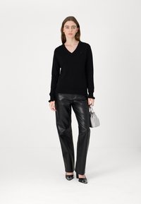 Schwarzer V-Ausschnitt-Pullover aus leichtem Strick, kombiniert mit hochgeschnittenen schwarzen Kunstlederhosen. Das Model hält eine kleine graue Handtasche. Schuhe mit glänzenden Akzenten.