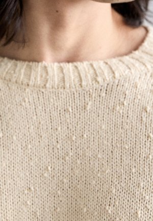 Gros plan sur le cou et les épaules d'une personne portant un pull en maille texturée crème avec un col rond côtelé.