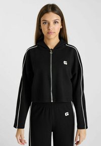 Zwarte zip-up hoodie met een cropped design, witte zijstrepen en logo op de borst. Zacht materiaal met een capuchon en een relaxte fit.