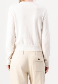 Bílý svetr s texturovaným povrchem, kulatým výstřihem a žebrovaným lemem, kombinovaný s béžovými kalhotami s boční kapsou a detaily na knoflík.