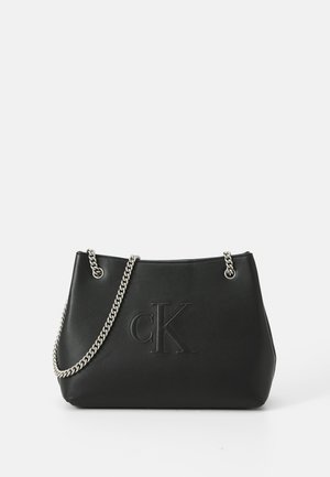 Sac à main en cuir noir avec logo "cK" embossé et bandoulière chaîne argentée sur fond blanc.