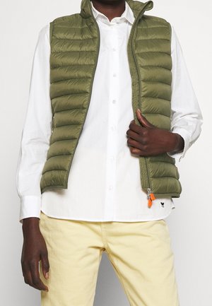 Olivgrüne gepolsterte Weste mit Reißverschluss, hohem Kragen und orangefarbener Reißverschlusslasche, getragen über einem weißen Hemd und gelben Hosen.