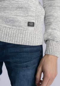 Maglione a maglia grigio chiaro con orlo e polsini a coste, dotato di un piccolo etichetta scura. Abbinato a jeans di denim blu scuro.