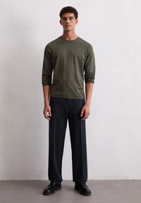 Mann steht gegen eine weiße Wand, trägt ein olivgrünes Langarmhemd, schwarze weit geschnittene Hosen und schwarze Schuhe, schaut nach vorne mit entspannten Armen.