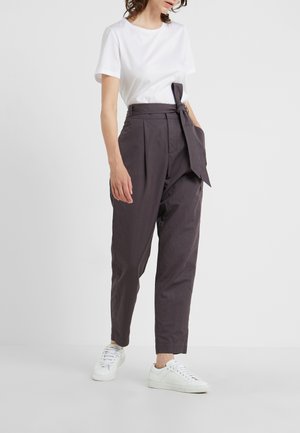 Kobieta w białej koszuli z krótkim rękawem, ciemnoszare spodnie paperbag z wysokim stanem i wiązaniem w pasie oraz białe trampki na jednolitym tle.