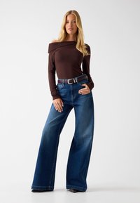 Top marrone a maniche lunghe con spalle scoperte abbinato a jeans blu a gamba larga e vita alta con cintura marrone, su sfondo bianco uniforme.