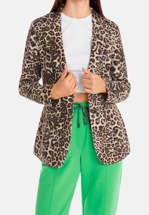 Personne portant un blazer à imprimé léopard sur un crop top blanc et un pantalon vert vif à cordon, sur un fond uni.