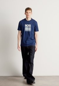 Temno modra majica s kratkimi rokavi z gradientnim logotipom "BOSS", kombinirana s temnimi kavbojkami. Model stoji pred enobarvnim ozadjem in prikazuje priložnostni videz.