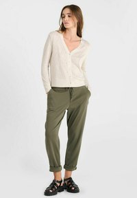 Hellbeige Cardigan mit V-Ausschnitt und Knöpfen auf der Vorderseite, kombiniert mit lockeren olivgrünen Hosen. Schwarze Sandalen mit Riemchen runden das Outfit ab.