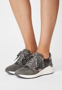 Šedé semišové tenisky s texturovanými akcenty, kontrastní bílou podrážkou a vzorovanými tkaničkami. Boční zip přidává funkčnost designu.