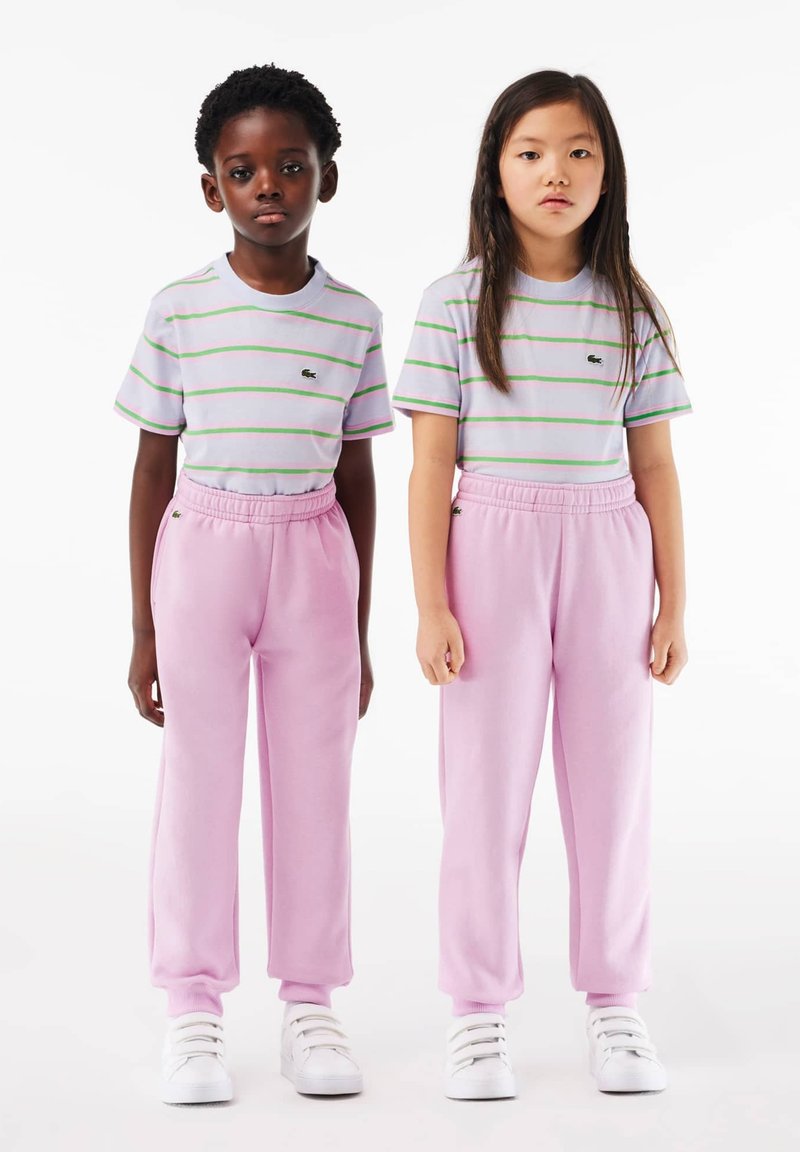 Deux enfants se tiennent côte à côte. Ils portent des t-shirts rayés violet clair et des pantalons de survêtement roses, complétés par des baskets blanches.