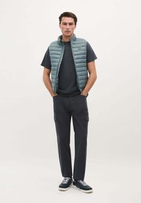 Lichtblauw gewatteerd vest over een marineblauwe t-shirt, gecombineerd met donkere cargobroeken en marineblauwe sneakers. Het vest heeft een hoge kraag en een ritssluiting aan de voorkant.