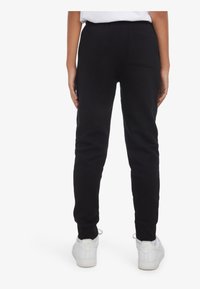 Schwarze Jogginghose mit figurbetontem Schnitt, elastischem Bund und einer Gesäßtasche. Kombiniert mit weißen Sportschuhen.