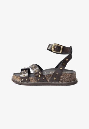 Sandalia de cuero marrón con tachuelas doradas, correa ajustable en el tobillo con hebilla dorada y suela gruesa de corcho moteado.