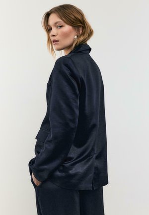 Junge Frau mit kurzen blonden Haaren, die einen dunkelblauen Blazer und passende Hose trägt, die Hände in den Taschen hat und über die Schulter blickt vor einfachem Hintergrund.