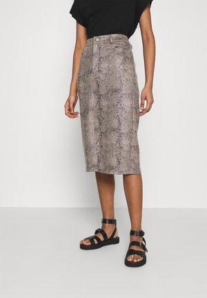 Kvinde iført en knælængde nederdel med slangehudsmønster, sort top og sorte buksesandaler, stående mod en hvid baggrund.