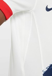 T-shirt de sport blanc avec une surface texturée, présentant des accents rouges sur les manches et un logo Nike bleu. Le design comprend des détails de panneaux.