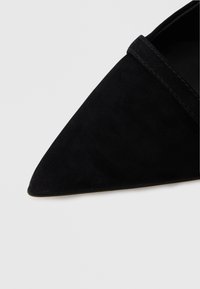 Svart semsket skinnsko med spiss tå, jevn tekstur og minimal sømdetalj langs kragen. Flat såle, uten synlig maskinvare.